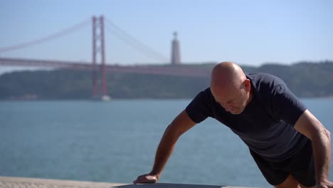 Bald-mature-sportsman-doing-push-ups-at-riverside